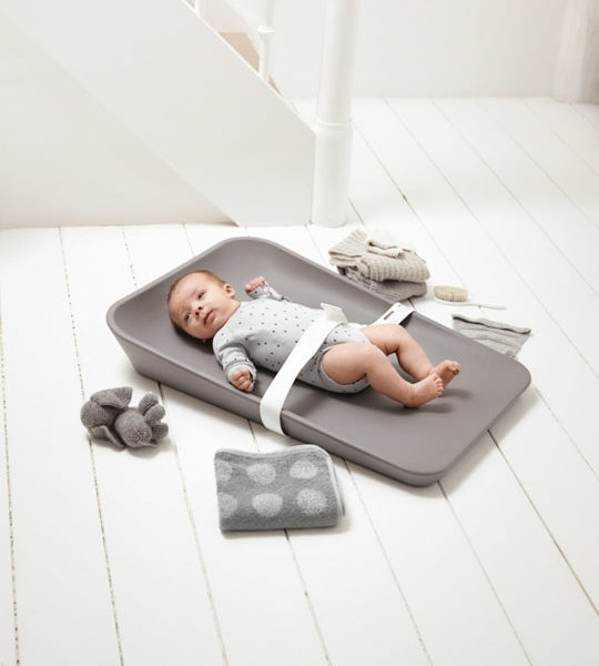 Baby strapped in and laying on grey changing mat on a white floor with grey towel, toy and clothes